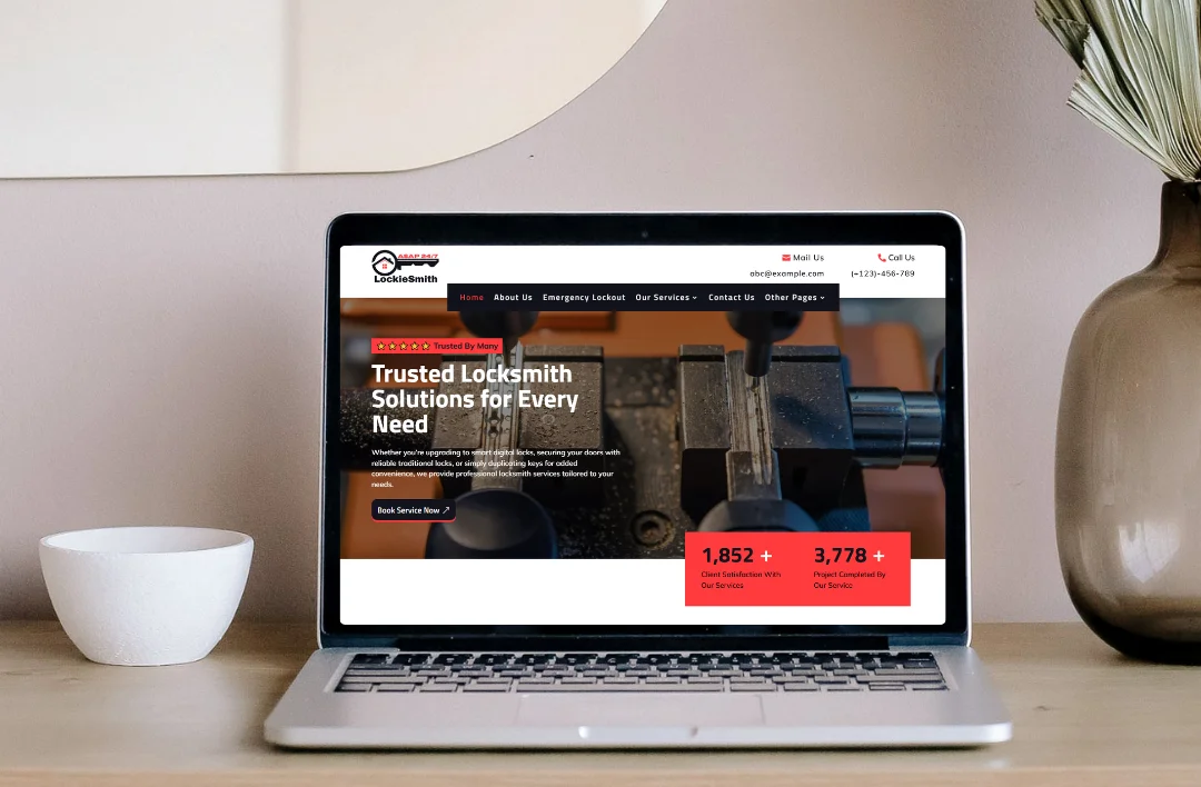 A silver laptop on a light wood desk displaying the homepage of "LockieSmith," a locksmith service website. The screen shows a red and black themed hero section with a locksmith at a key-cutting machine and a headline that reads "Trusted Locksmith Solutions for Every Need." Statistical counters for client satisfaction and completed projects are highlighted in red at the bottom. The laptop is placed next to a small white bowl and a brown vase with dried plants.