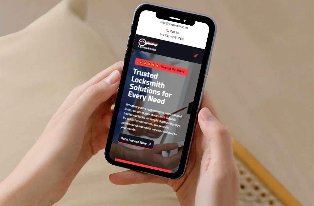 A close-up shot of a person's hands holding a smartphone, displaying the mobile-responsive version of the "LockieSmith" website. The screen shows a professional red and black theme with a prominent headline, "Trusted Locksmith Solutions for Every Need," and a "Book Service Now" button. The layout demonstrates easy navigation and clear contact information for mobile users needing emergency locksmith services.