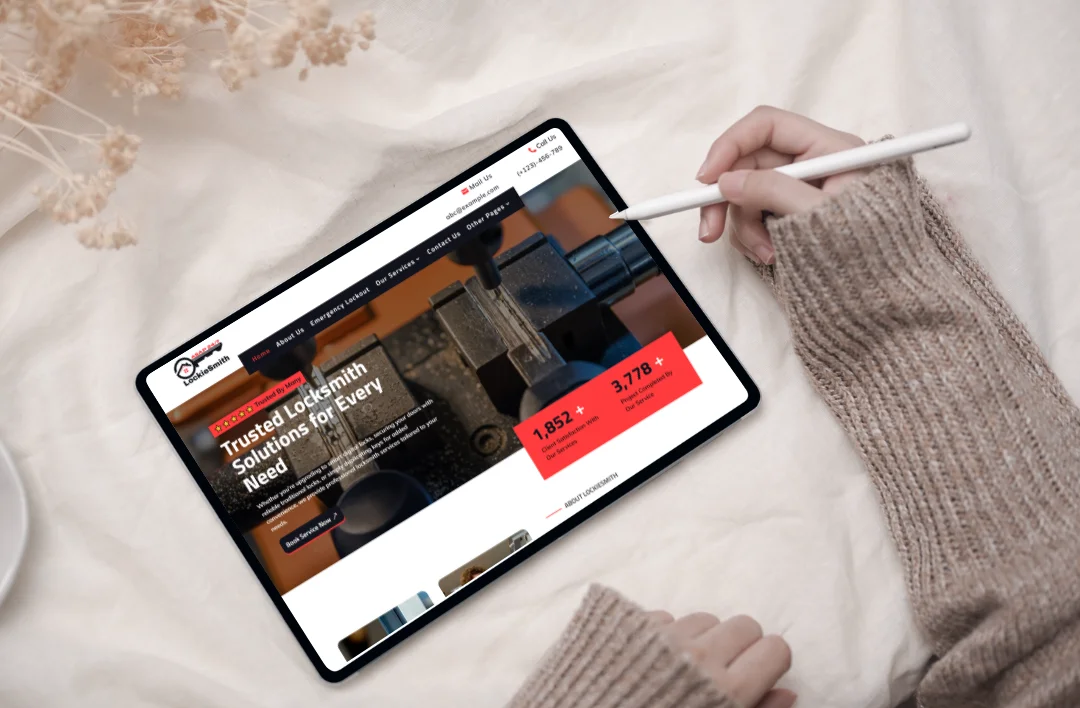 An overhead view of a person using a white stylus on a tablet that displays the "LockieSmith" locksmith website. The screen shows a professional red, black, and white theme with a headline for trusted locksmith solutions and large red call-out boxes for client satisfaction and project statistics. The tablet is placed on a soft white surface decorated with dried flowers.