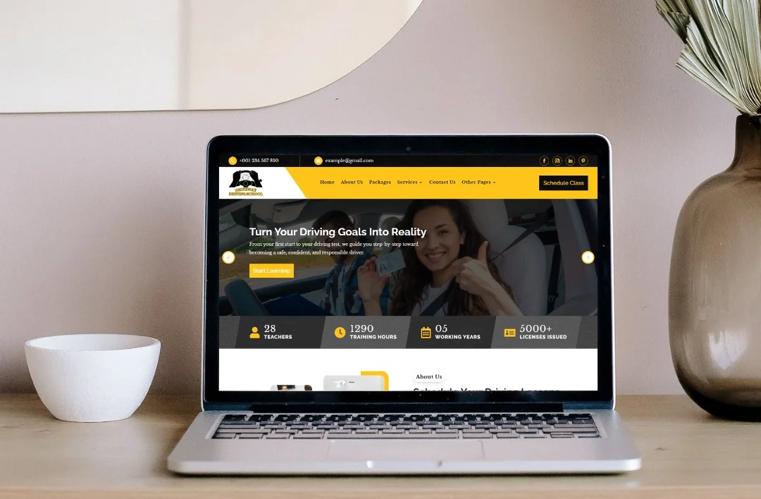 A silver laptop on a wooden desk displaying the homepage of a driving school website. The screen features a yellow and black theme with a headline that reads "Turn Your Driving Goals Into Reality" and a banner showing key statistics such as "5000+ Licenses Issued." Beside the laptop sits a small white bowl and a brown glass vase with dried botanical stems against a neutral-toned wall.