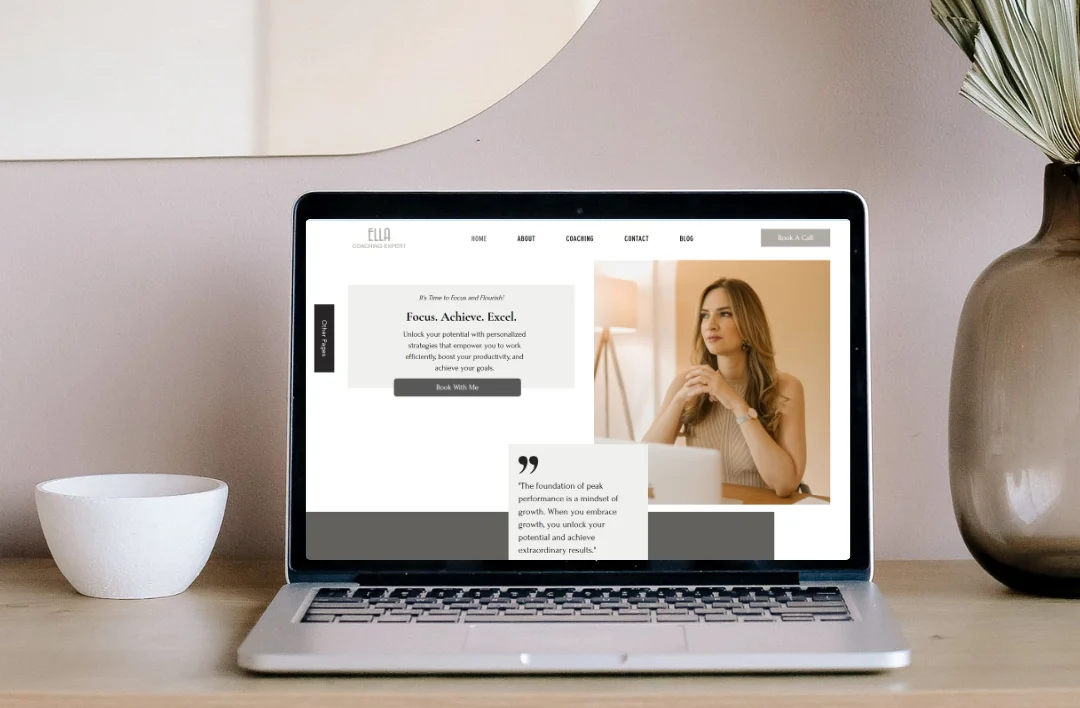 Minimalist lifestyle mockup of a 'Productivity Coach' website template displayed on a laptop screen. The laptop sits on a clean wooden desk next to a white ceramic bowl and a glass vase with dried plants. Elegant and professional aesthetic.