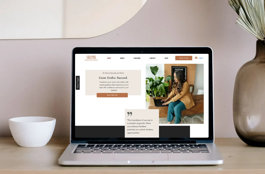 A professional laptop mockup of a Business Coach website template on a light wooden desk. The screen displays a warm earth-tone and minimalist white design with a professional woman sitting at a desk. Styled with a white bowl and a glass vase with dried pampas grass in a bright, modern room.