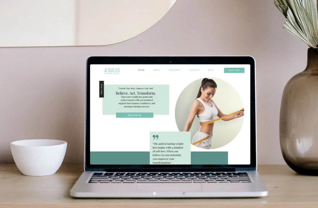 Professional lifestyle mockup of a 'Weight Loss Coach' website template displayed on a laptop screen. The laptop is placed on a light wooden desk in a bright, modern room next to a white bowl and a glass vase with dried pampas grass. Clean mint green and white design.