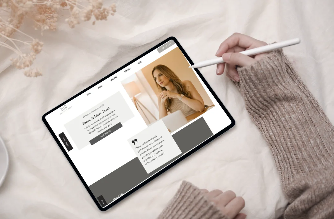 Top-down lifestyle mockup of a 'Productivity Coach' Wix website template displayed on a tablet with a stylus. The minimalist design features a professional portrait and a clean white-and-grey layout. Surrounded by soft fabric and dried flowers for an elegant aesthetic.