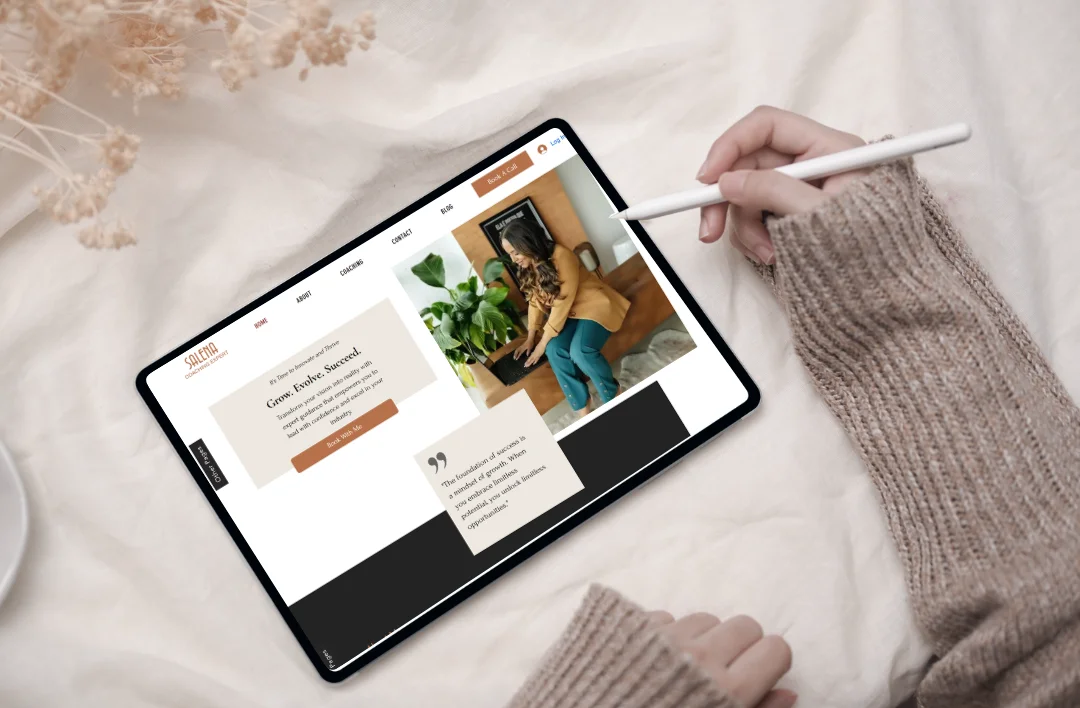 A top-down lifestyle mockup of the 'Selena' Business Coach Wix website template displayed on a tablet with a stylus. The screen features a warm earth-tone (terracotta and beige) minimalist design with a professional woman working at a laptop. Styled on a soft white fabric background with dried flowers.